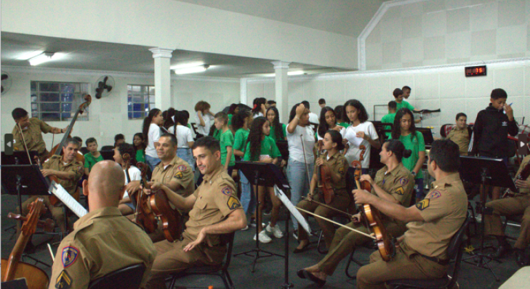 #paratodosverem: a foto mostra uma sala ampla, com teto alto e paredes claras, onde vários(as) policiais militares e estudantes estão reunidos(as). Em primeiro plano, há um grupo de policiais fardados(as) sentados(as) em cadeiras, segurando instrumentos de cordas, como violinos, violas e contrabaixos. Ao fundo, há estudantes uniformizados(as), muitos(as) em pé, segurando folhas de papel. 