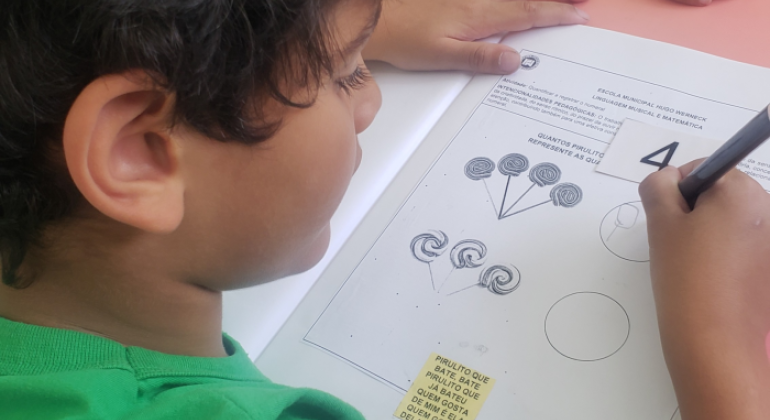 #paratodosverem: na foto, uma criança com camiseta verde realiza uma atividade escolar, que consiste em contar pirulitos desenhados e escrever o número correspondente. A criança segura um lápis e está prestes a escrever. No canto inferior da folha, há a letra da cantiga "Pirulito que bate, bate / Pirulito que já bateu / Quem gosta dela sou eu!"