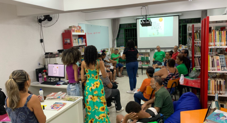 #paratodosverem: na imagem, pessoas adultas participam de uma roda de conversa em uma biblioteca escolar. A maioria está sentada em cadeiras e pufes coloridos. Ao fundo, uma mulher de camiseta preta conduz a atividade, com um slide projetado no quadro em que está escrito “O que você faz com uma ideia?” Estantes com livros, mesas e um computador compõem o ambiente. Algumas pessoas de pé acompanham a conversa.