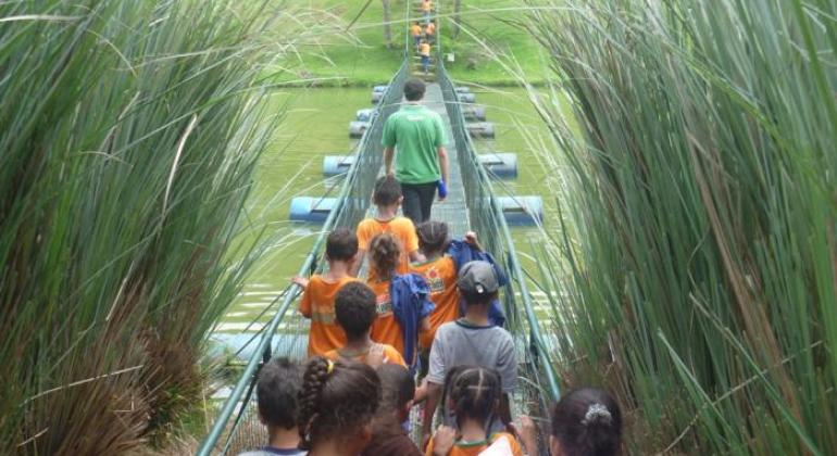 #paratodosverem: crianças com camisetas laranja e cinza atravessam uma ponte estreita sobre um lago, acompanhadas por um adulto. A ponte flutua com apoio de tambores azuis e está cercada por vegetação alta. A cena transmite aventura, natureza e aprendizado ao ar livre, representando uma atividade do projeto Aulas Passeio.