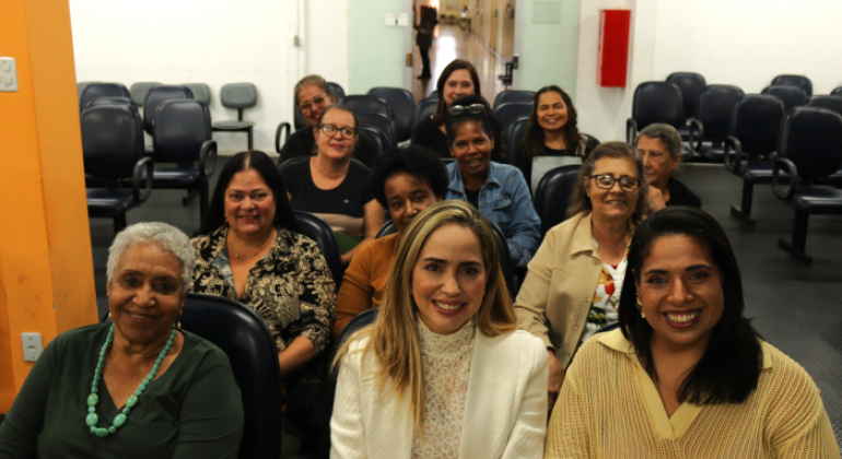 #paratodosverem: na imagem, doze mulheres estão reunidas em um auditório, sentadas em cadeiras de auditório e sorrindo. O auditório tem paredes brancas, cadeiras escuras e uma porta de vidro, ao fundo, decorada com bandeirinhas coloridas. 