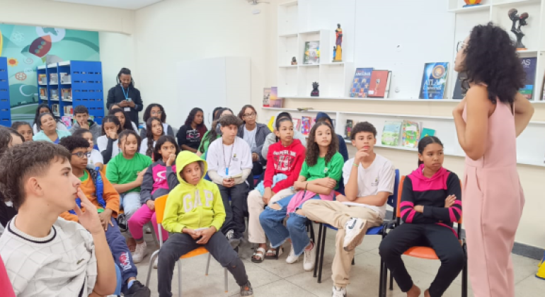 #paratodosverem: a imagem mostra uma roda de conversa em uma biblioteca. Um grupo de crianças e adolescentes está sentado, prestando atenção em uma mulher que fala de pé, à frente do grupo. Ao fundo, há estantes com livros, cartazes e decorações. 