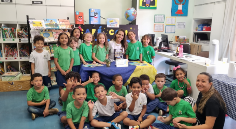 #paratodosverem: foto de uma turma de crianças pequenas uniformizadas em uma biblioteca. Elas estão reunidas em volta de uma mesa decorada com tecido azul e amarelo, acompanhadas por duas educadoras. No fundo, há prateleiras com livros e desenhos coloridos nas paredes.