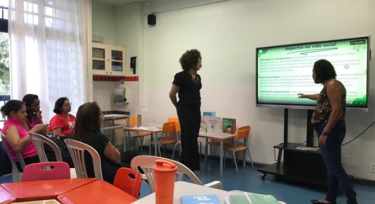 #paratodosverem: na foto, há quatro mulheres, à esquerda, sentadas em cadeiras plásticas brancas, enquanto duas estão de pé, à frente da sala. Uma destas aponta para uma tela interativa onde está sendo exibido um slide intitulado "Impactos na vida social". A sala tem paredes e cortinas brancas, armários com portas de vidro, além de mesas e cadeiras nas cores laranja, azul e vermelha.