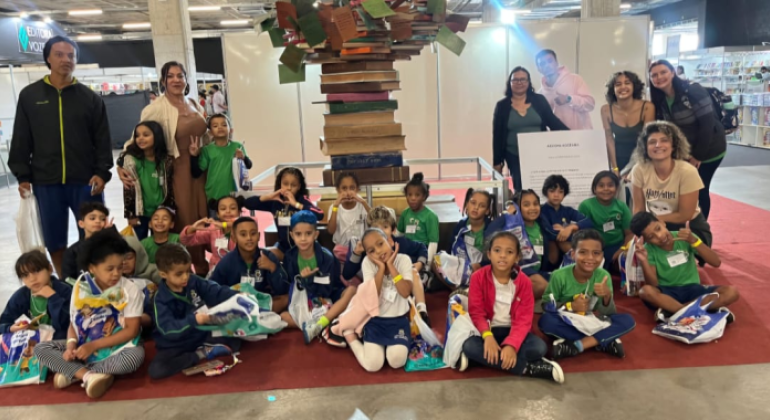 #paratodosverem: um grupo de crianças e pessoas adultas posa para foto, em frente a uma escultura feita com livros empilhados. As crianças estão sentadas no chão, com sacolas nas mãos, sorrindo. Ao fundo, há estandes de livros e uma placa com texto explicativo. O local tem piso de concreto e iluminação artificial.