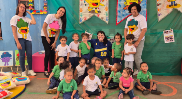 #paratodosverem: A foto mostra um grupo de crianças pequenas sentadas e em pé no pátio escolar, acompanhadas por quatro mulheres. O fundo é decorado com bandeiras coloridas ilustradas com personagens sorridentes e os nomes das turmas, como “Turma do Martelo” e “Turma do Trenzinho”. Há também trabalhos manuais, desenhos e almofadas estampadas com as mesmas figuras. Em meio as crianças, agachada com uma blusa azul, está a artista Raquel Bolinho.