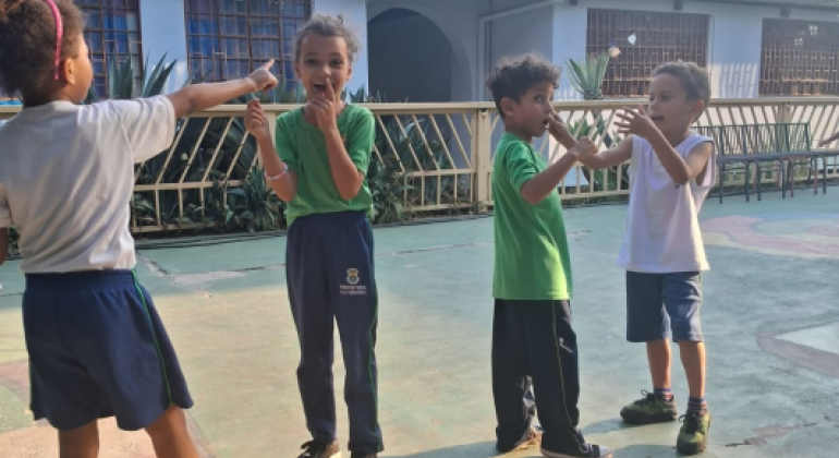 Brincadeiras animam os estudantes #paratodosverem: Duas meninas e dois meninos brincam em um pátio de cimento. A menina de costas aponta para a outra, enquanto as outras três crianças, incluindo um menino de uniforme verde e um de uniforme branco, riem e interagem com expressões animadas.