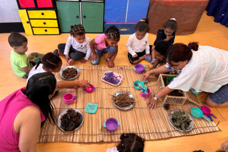 #paratodosverem: Em uma sala, crianças e duas professoras estão sentadas no chão de madeira, em volta de um tapete de palha. Sobre o tapete, há bandejas com elementos da natureza, como pinhas e folhas, que as crianças usam para brincar. A cena, vista de cima, mostra o foco na exploração sensorial e no aprendizado lúdico.
