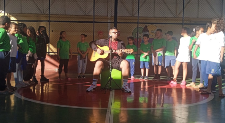 #paratodosverem: em uma quadra, há 15 crianças de pé, dispostas em semicírculo, para apresentação musical. No centro da quadra, um adulto está sentado em um pufe verde, tocando violão com um microfone à sua frente. Os(As) estudantes usam uniformes verdes ou brancos com calças azuis; já o adulto veste uma blusa vermelha, jaqueta e bermuda pretas. 