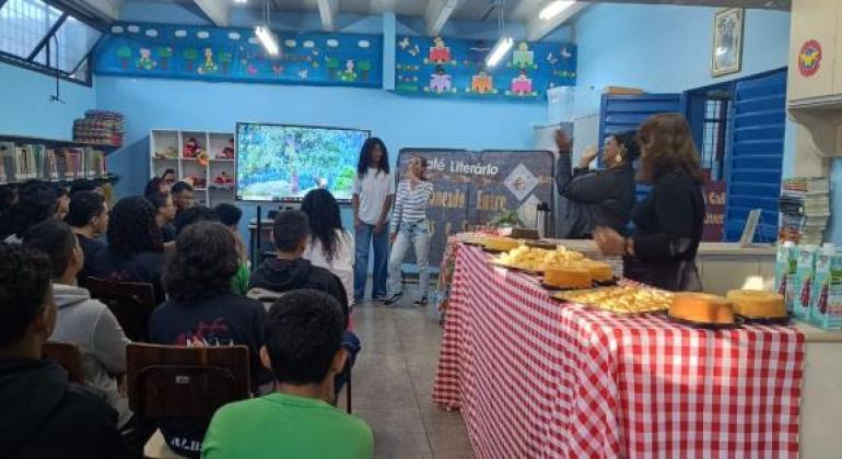 #paratodosverem: a foto mostra o interior de uma biblioteca decorada para o “Café Literário”. Vários(as) estudantes estão sentados(as) de frente para um telão e duas alunas estão, à frente do grupo, apresentando. À direita da imagem, há uma mesa, coberta com toalha xadrez vermelha e branca, repleta de alimentos como bolos, pães de queijo e sucos. Atrás da mesa, duas mulheres assistem às estudantes.