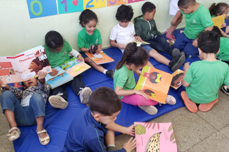 O projeto visa a fortalecer a identidade das crianças #paratodosverem: Um grupo de crianças, a maioria de uniforme verde, está confortavelmente sentado ou deitado em um tapete azul no chão da sala de aula. Cada criança segura um livro, absorvidas pela leitura, algumas sozinhas e outras próximas.