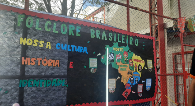 #paratodosverem Painel com o título “Folclore Brasileiro: nossa cultura, história e identidade”, decorado com letras coloridas. No centro, um mapa do Brasil com figuras de personagens folclóricos distribuídos pelas regiões. O cartaz está preso em uma grade de ferro, com bandeirinhas de festa junina ao redor.