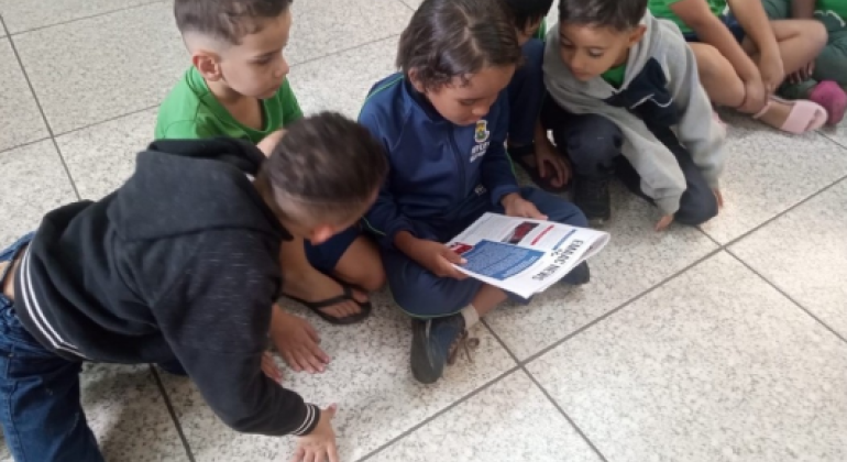 #paratodosverem: Quatro meninos estão sentados no chão de um ambiente escolar. Eles se reúnem em volta de um pequeno jornal ou revista, lendo atentamente. A cena, que mostra o grupo inclinado e focado na leitura, transmite a ideia de um momento de aprendizado e curiosidade compartilhado entre os amigos.