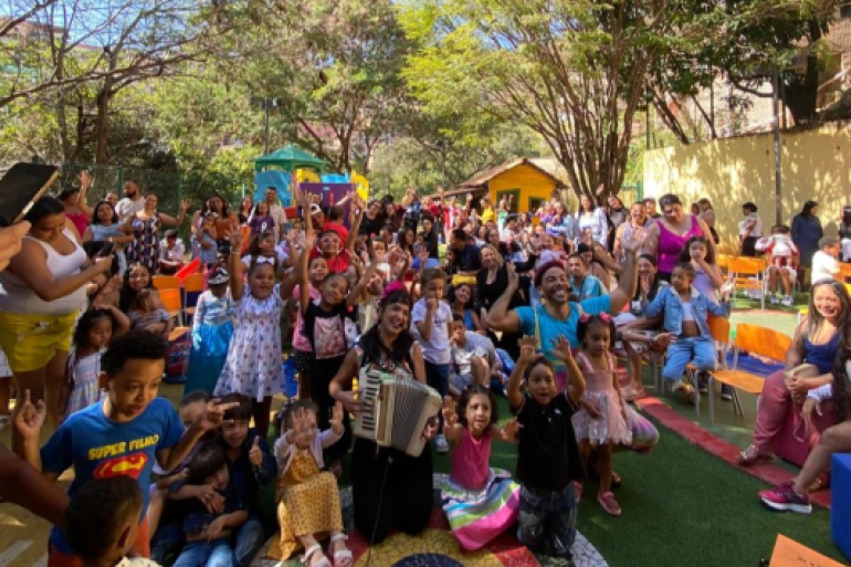 #paratodosverem: em um pátio escolar ao ar livre, um grande grupo de crianças e adultos participa de um momento de confraternização. No centro, uma artista toca sanfona e sorri, rodeada por crianças que levantam as mãos animadas. Ao lado dela, outro artista interage com o público, também sorridente. Muitas famílias estão sentadas e em pé ao fundo, acompanhando a atividade.