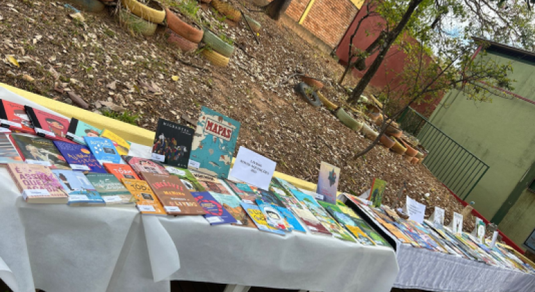 #paratodosverem: Em mesas cobertas com toalhas brancas, vários livros coloridos estão expostos ao ar livre, em frente a um jardim com pneus pintados usados como floreiras. As capas chamam a atenção pela diversidade de temas e cores.