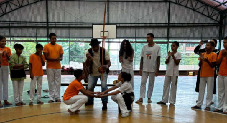 Estudantes celebram a capoeira em evento. #paratodosverem: Um grupo de jovens, vestindo uniformes de cores laranja e branco, está disposto em um semicírculo em um ginásio coberto. No centro, dois participantes estão agachados, tocando as mãos, em um ato de capoeira. Ao fundo, um homem toca um berimbau. A cena ocorre sob um telhado arqueado de metal, com uma tabela de basquete visível.