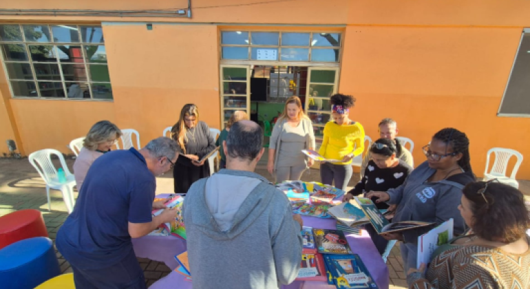#paratodosverem: Um grupo de adultos está ao redor de uma mesa coberta com toalha roxa, onde há vários livros coloridos expostos. Algumas pessoas folheiam os livros, enquanto outras observam. Ao fundo, vê-se uma parede laranja com janelas de vidro e cadeiras plásticas brancas encostadas.