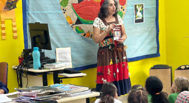 #paratodosverem: a foto mostra uma mulher de vestido colorido e estampado exibindo um livro para um grupo de crianças uniformizadas sentadas no chão de uma sala de aula. Ela está posicionada em frente a um painel azul com um desenho circular ao centro, contendo diversas frutas. Ao lado, há uma mesa com pilhas de livros e outra com um computador. 