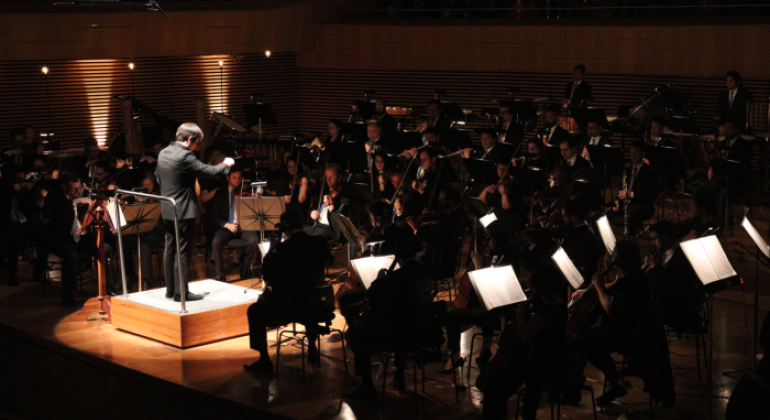 #paratodosverem: a imagem mostra a Orquestra durante a apresentação na Sala Minas Gerais. O maestro está em pé, ao centro, em uma pequena estrutura elevada, regendo, com movimentos das mãos, músicos e musicistas, que estão organizados(as) no palco, conforme os instrumentos: cordas, à frente; sopros, ao centro, e percussão, ao fundo. Luzes incidem sobre o maestro e sobre as partituras. 
