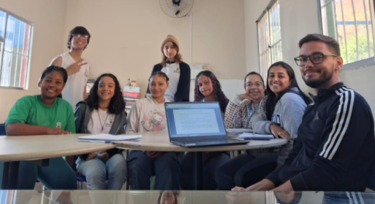 #paratodosverem: Grupo de jovens e um adulto estão sentados e em pé ao redor de uma mesa em sala iluminada, com janelas laterais e um ventilador. Sobre a mesa há um notebook aberto e cadernos. Todos sorriem para a foto, alguns usando moletom, outros camiseta, e uma jovem de touca marrom está em pé ao fundo.