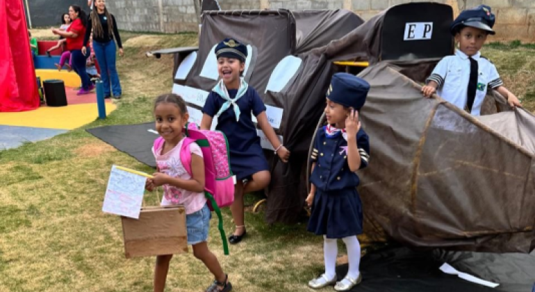 #paratodosverem: Em um jardim, crianças brincam com um avião de papelão feito por elas mesmas. Duas meninas e um menino estão fantasiados de pilotos e tripulação, sorrindo e se divertindo. Uma das garotas, com uma mochila rosa nas costas e um papel nas mãos, olha diretamente para a câmera. A cena transmite a alegria de uma brincadeira cheia de imaginação.