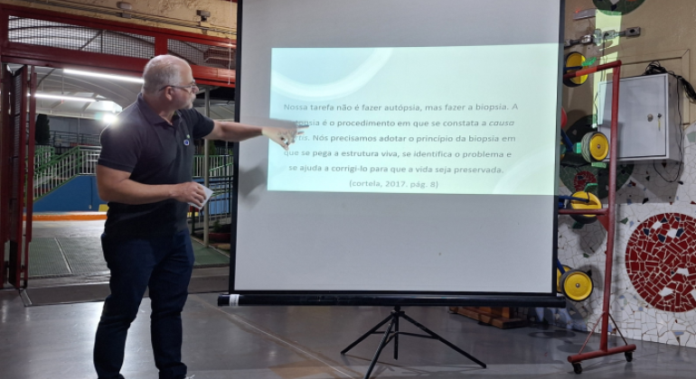 #paratodosverem: um homem de cabelo grisalho, usando camiseta preta e calça jeans, aponta para um slide projetado em uma tela branca. A apresentação acontece em um espaço escolar, com piso de cimento e paredes decoradas com mosaicos coloridos.