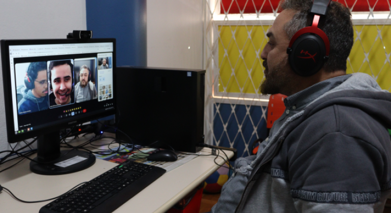 #paratodosverem: na foto, o professor está sentado em frente a um computador participa de uma videoconferência com três outros estudantes, visíveis na tela. Ele usa fones de ouvido vermelhos e pretos e veste um casaco cinza com capuz. O ambiente tem paredes coloridas e uma janela com grade branca ao fundo.