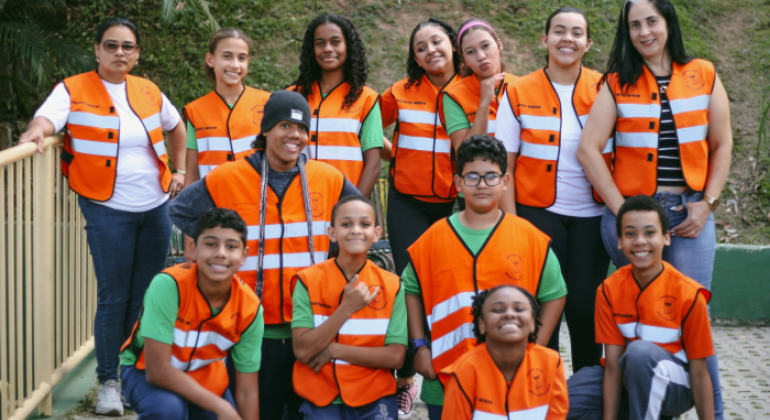 #paratodosverem: a foto mostra um grupo de estudantes com duas educadoras. Todos(as) vestem colete laranja com faixas refletivas brancas e detalhes pretos sobre a blusa. Eles(as) estão ao ar livre e, ao fundo, há um gramado. 