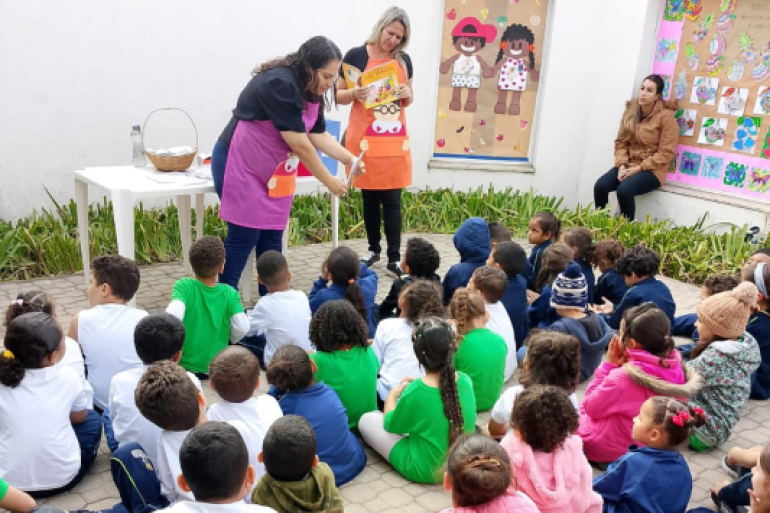 #paratodosverem: Em uma sala de aula, um grupo de adultos está em pé, formando um círculo. Eles seguram fios de barbante, criando uma teia entre si, como parte de uma atividade coletiva. Ao fundo, há cadeiras laranjas encostadas na parede, armários coloridos e materiais pedagógicos. A sala é iluminada por janelas e há um ventilador na parede.
