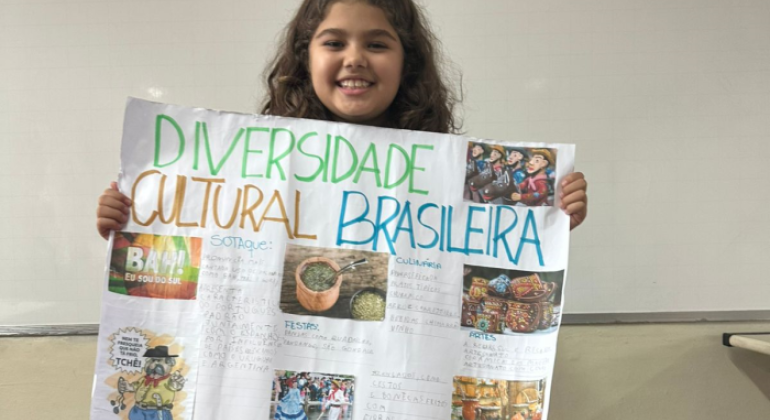 #paratodosverem: na imagem, uma menina sorridente segura um cartaz colorido com o título "Diversidade Cultural Brasileira", que mostra imagens e textos sobre sotaques, culinária, festas e artes do Brasil. Ao fundo, há um quadro branco de sala de aula.