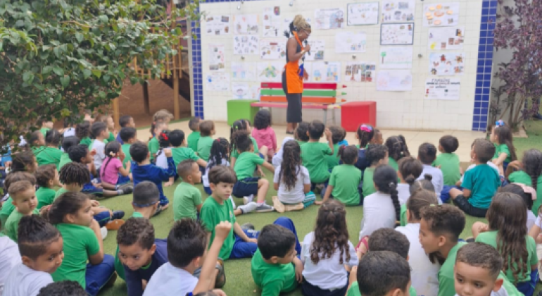 #paratodosverem: Crianças de camiseta verde estão sentadas na grama, em uma grande roda, assistindo a uma contadora de histórias ou professora. A mulher, de blusa preta e avental laranja, está em pé em frente a um mural cheio de desenhos e recados.