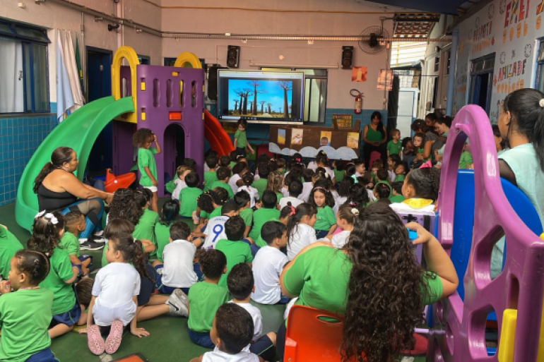 Programação especial envolveu a árvore Baobá #paratodosverem: Em uma sala de aula colorida, com um grande brinquedo infantil, várias crianças de camiseta verde estão sentadas no chão em semicírculo, voltadas para uma tela digital. A tela exibe imagens de árvores. Duas professoras estão com o grupo. A cena, vibrante e cheia de cores, mostra um momento de aula interativa e lúdica.
