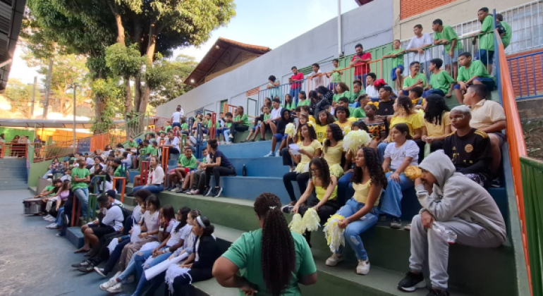 #paratodosverem: na fotografia, estudantes estão sentados(as) em arquibancadas coloridas ao ar livre, eles(as) estão olhando para frente. Muitos(as) vestem camisetas verdes, brancas ou amarelas, e alguns/algumas seguram pompons. Há árvores ao fundo, além de construções escolares.