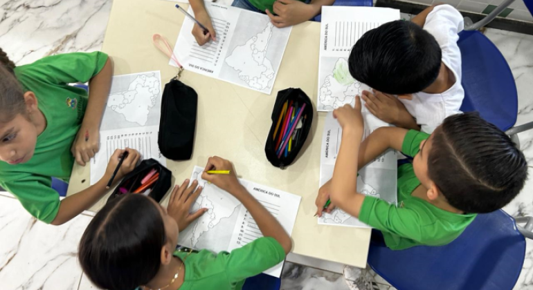 A imagem mostra um grupo de cinco crianças reunidas em torno de uma mesa em uma sala de aula. Elas estão vestindo camisetas verdes, com exceção de uma criança que veste branco, como parte de um uniforme escolar. As crianças estão realizando uma atividade educacional, escrevendo e colorindo com lápis de cor e lápis grafite. Elas parecem concentradas e envolvidas na atividade, compartilhando materiais como estojos e lápis. O ambiente possui cadeiras azuis, uma mesa bege e um piso de mármore branco com veios e