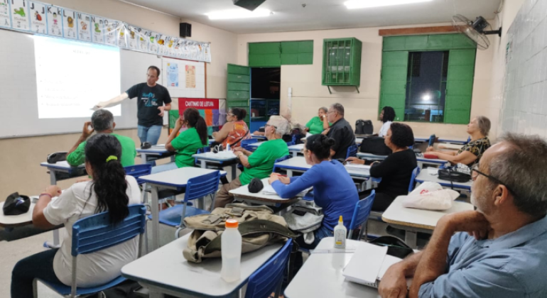 #paratodosverem: a foto mostra um educador na frente de um quadro, em uma sala pintada de bege, com janelas e portas verdes. Acima do quadro, há papéis com as letras do alfabeto. No teto, um projetor exibe uma apresentação. As carteiras estão quase todas ocupadas com pessoas adultas de diversas idades. Algumas usam uniformes e estão prestando atenção no que o professor explica.