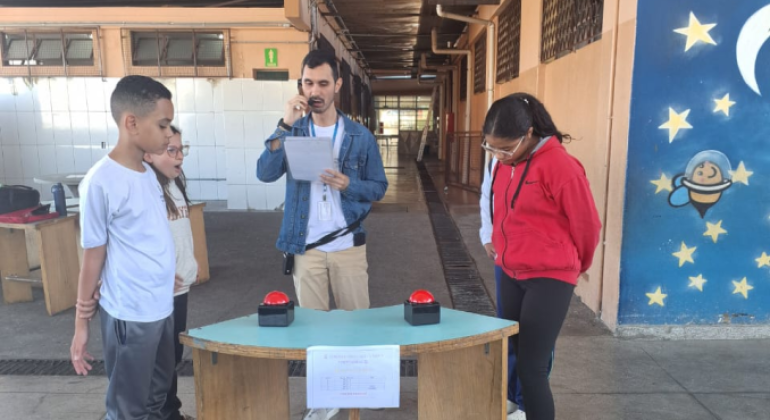 #paratodosverem: Na foto, dois estudantes participam de um jogo de perguntas e respostas em uma escola, enquanto um professor conduz a atividade com um microfone e uma folha de anotações. À frente deles há uma mesa com botões vermelhos, simulando um quiz. O clima é de concentração e aprendizado em um ambiente descontraído e educativo.