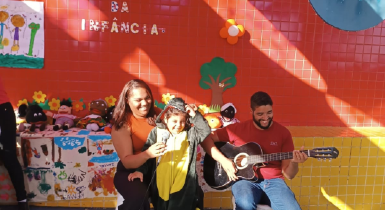 #paratodosverem: em frente a um painel laranja decorado com flores e brinquedos, uma criança fantasiada de dinossauro canta sorridente ao microfone, acompanhada por uma mulher ao lado e um homem tocando violão. A cena faz parte da “Semana da Infância”.