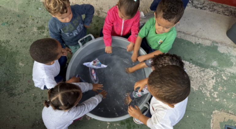 #paratodosverem: a foto mostra crianças pequenas ao redor de uma grande bacia cheia de água, brincando com barquinhos feitos de papel. Elas observam, atentas, os barquinhos boiando, enquanto algumas colocam as mãos na água. 