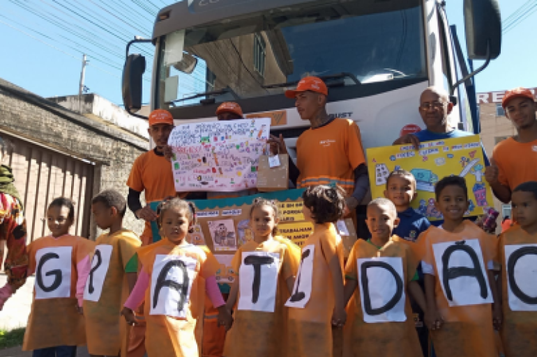 #paratodosverem: Um grupo de crianças veste aventais laranjas com letras que formam a palavra “GRATIDÃO”, posando em frente a um caminhão branco. Atrás delas, garis uniformizados de laranja seguram cartazes coloridos com mensagens de agradecimento e desenhos. A cena acontece em uma rua, com céu azul e fiação elétrica visível ao fundo.