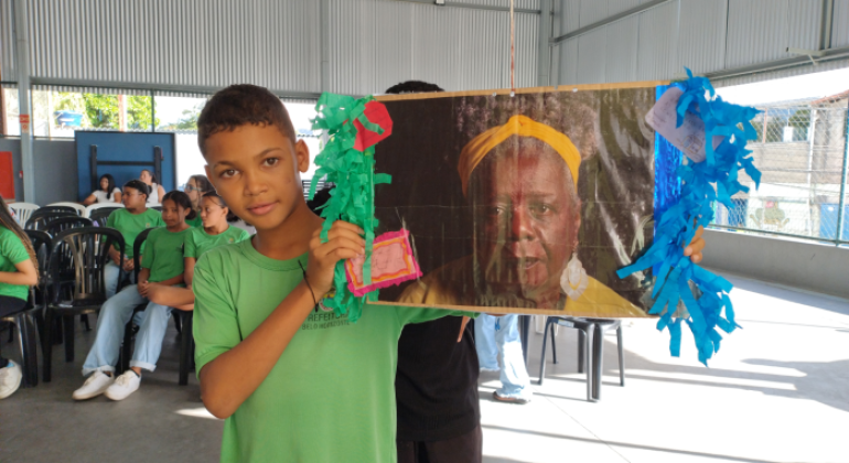 #paratodosverem: a imagem mostra, em um espaço coberto e fechado nas laterais por grades, um menino com uniforme escolar verde segurando; à sua direita, uma foto impressa de Conceição Evaristo colada em um cartaz com papel crepom verde e azul. Ao fundo, outros(as) estudantes uniformizados(as) estão sentados(as) em cadeiras de plástico brancas.