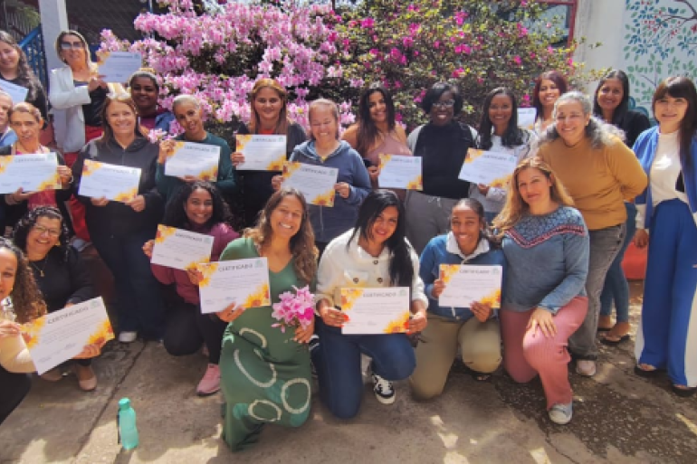 Encontro promove momento de troca de ideias e afetividade #paratodosverem: Um grupo de mulheres posa sorridente, ao ar livre, segurando certificados com bordas floridas. Atrás delas, há arbustos floridos de cor rosa e uma parede colorida com desenhos de animais. Algumas estão agachadas na frente, enquanto outras permanecem em pé, formando uma grande fileira.