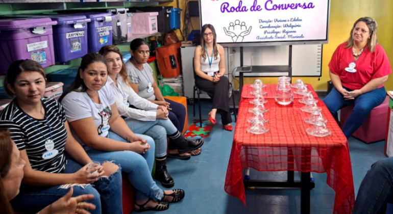 #paratodosverem: a foto mostra uma roda de conversa realizada em uma sala com parede amarela e uma estante com caixas organizadoras, à esquerda. Em frente à estante, algumas mulheres estão sentadas. No centro da sala, há uma mesa coberta com toalha vermelha, com várias xícaras de vidro e uma jarra com água. Ao fundo, um monitor exibe a arte gráfica do evento com o título “Roda de Conversa”.