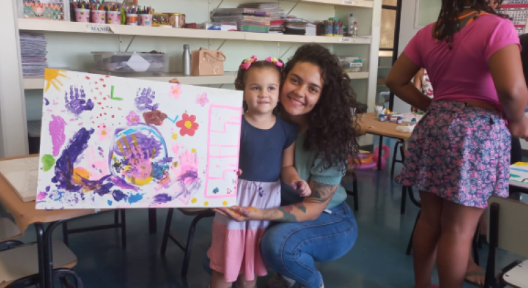 #paratodosverem: na imagem, em primeiro plano, mãe e filha estão em uma sala com paredes brancas e chão verde musgo. A mãe está agachada, à altura da criança, vestida com calça jeans azul e blusa verde. A criança usa um vestido azul marinho, branco e rosa. A mãe segura um quadro multicolorido pintado pela filha, com a presença de flores e mãos. Ao fundo, há prateleiras brancas com materiais pedagógicos, como papéis e recipientes cheios de lápis de cor. À direita, estão duas mulheres adultas.