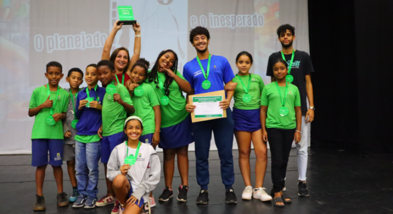 #paratodosverem: na imagem, um grupo diverso de estudantes e educadores(as), usando camisetas verdes e medalhas, posa sorridente no palco com certificado e troféu do VIII FEIMC-BH 2024. Ao fundo, há um telão com o tema do evento: “O Planejado e o Inesperado”. A imagem expressa celebração, orgulho e união após a premiação.