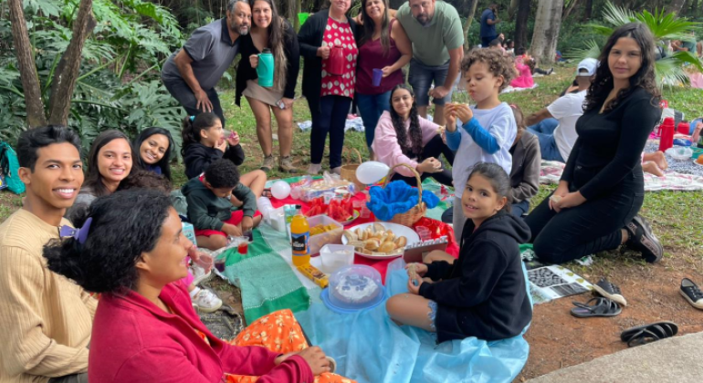 #paratodosverem: a imagem mostra, em um parque, um grupo de pessoas, incluindo pessoas adultas e crianças, reunido num piquenique em meio à natureza. Todos(as) estão em clima de alegria e descontração, alguns(mas) sentados(as) ao redor de uma toalha, sobre a qual há diversas guloseimas e bebidas, enquanto outros(as) estão em pé, ao fundo, sorrindo para a foto. O ambiente é arborizado, com bastante vegetação.