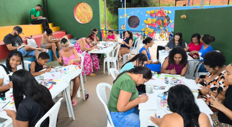 #paratodosverem: mulheres, crianças e alguns homens participam de uma atividade coletiva de pintura em telas, com mesas cheias de tintas, pincéis e telas, em um ambiente colorido, com a parte superior das paredes aberta ao exterior. Ao fundo da imagem, há brinquedos em uma área externa e, atrás deles, um muro azul com desenhos. Mais além, está uma área com plantas. 