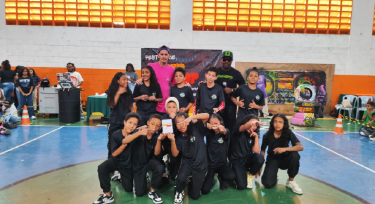 #paratodosverem: Um grupo de crianças e adolescentes veste roupas pretas iguais e posa no centro de uma quadra esportiva, alguns agachados e outros em pé, fazendo gestos com as mãos. Atrás deles, dois adultos estão de pé, um de rosa e outro de boné verde com microfone. No fundo há um banner “Festival Hip Hop”, grafite colorido e pessoas assistindo. Alguns estudantes na frente seguram um prêmio em formato de um retângulo branco.#paratodosverem: Um grupo de crianças e adolescentes veste roupas pretas iguais e