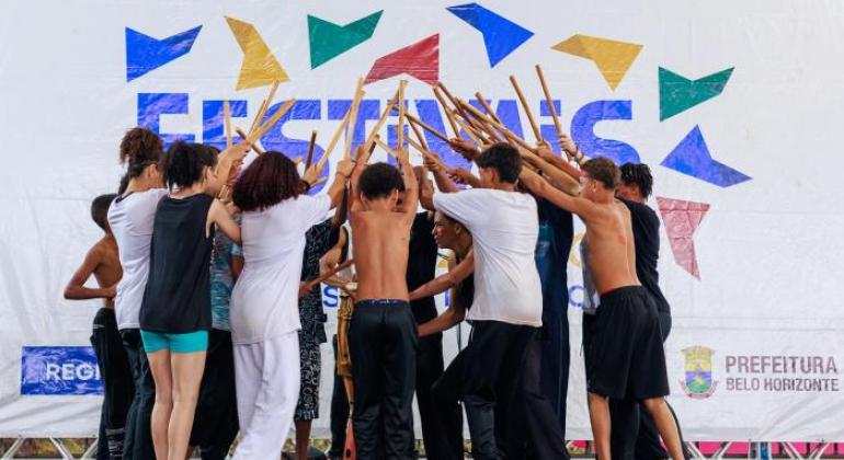 #paratodosverem: grupo de crianças e adolescentes se apresenta com bastões em frente a um painel com a palavra “FESTIVAIS”. Eles usam roupas brancas com camisetas coloridas e estão em formação. Ao fundo, adultos tocam tambores e pandeiro. A cena transmite energia, cultura e integração em um evento escolar.