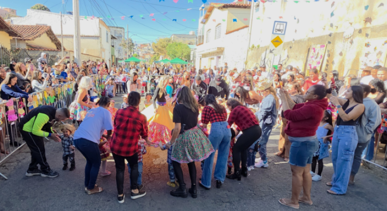 #paratodosverem: na foto, pessoas adultas caracterizadas para uma festa junina, em roda, seguram a barra do vestido de uma garota que está no centro, em uma rua enfeitada com bandeirinhas coloridas. Ao redor, familiares observam e registram o momento com celular. O céu está azul e o dia ensolarado. Grades decoradas com temas juninos cercam o espaço.