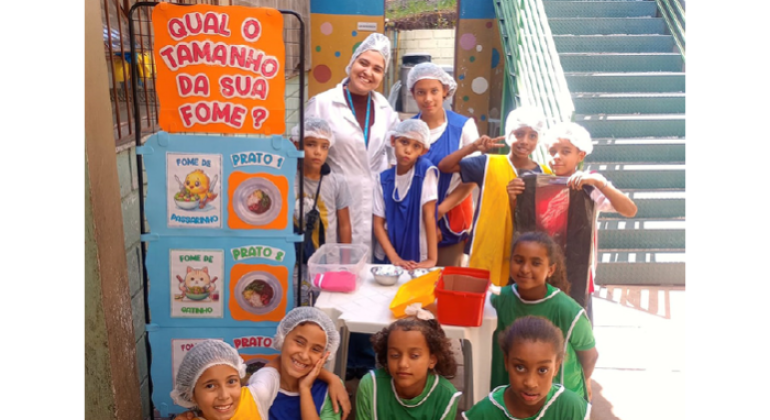 #paratodosverem: na foto, crianças posa ao lado de uma profissional de jaleco branco. À esquerda, um painel colorido exibe a pergunta: “Qual o tamanho da sua fome?” As crianças estão organizadas junto a uma mesa com caixas plásticas e bacias de metal. Algumas usam coletes coloridos e toucas. Ao fundo, vê-se uma escada verde e parte da área externa da escola.